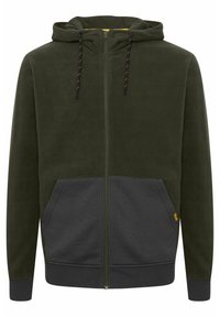 Sudadera con cremallera de color verde oliva con una capucha forrada en amarillo, bolsillos frontales de color gris oscuro y cordones en contraste. Textura de felpa suave.