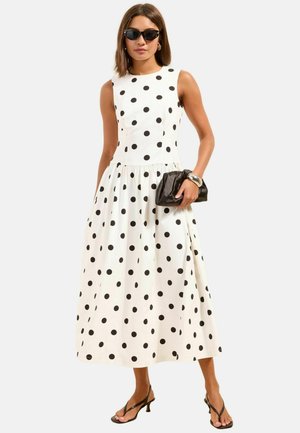 Žena oblečená v bielych šatách bez rukávov s čiernymi bodkami, čierne slnečné okuliare, drží čiernu kabelku a nosí čierne sandále na podpätku.