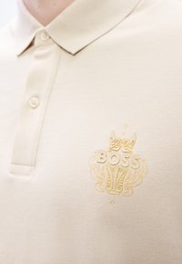Μπεζ πόλο πουκάμισο από υφασμάτινη υφή με χρυσό κεντητό λογότυπο "BOSS" και σχεδιασμό κορώνας στην αριστερή πλευρά του στήθους. Γιακάς με δύο κουμπιά.