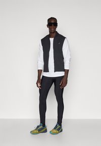Gilet noir sur une chemise à manches longues blanche, leggings noirs et chaussures de course vertes avec des accents bleus. Des lunettes de soleil complètent la tenue.