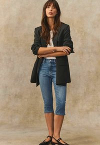 Svart randig oversized blazer, vit åtsittande t-shirt, ljusblå cropped jeans och svarta ballerinaskor. Modellen poserar mot en strukturerad beige bakgrund.