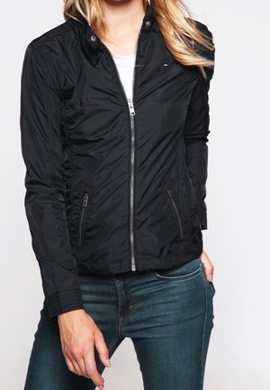 Frau trägt eine eng anliegende schwarze Jacke mit Reißverschluss und Reißverschlusstaschen, kombiniert mit blauen Denim-Jeans, die am Hals und an den Hüften gekürzt sind.