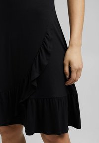 Main d'une femme reposant à côté d'une robe noire avec un ourlet à volants sur un fond gris uni.