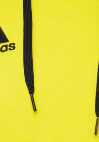 Sweat-shirt jaune avec cordons noirs, arborant un logo sur le côté gauche. Le tissu semble doux et texturé.