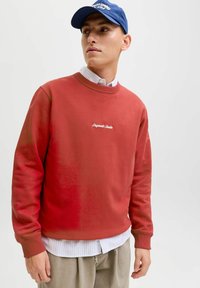 Czerwony sweter z okrągłym dekoltem, miękka tekstura, z białym haftowanym logo. Połączony w paskowanym koszulą i beżowymi spodniami. Niebieska czapka na głowie.