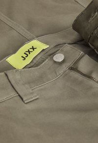 Olivgröna byxor med en gul etikett med "JJXX", en silverfärgad knapp och en dragkedjeficka, som framhäver slitstarkt tyg och rena sömmar.