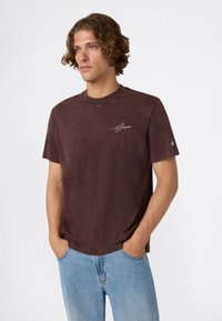 Joven con pelo rizado que lleva una camiseta de manga corta marrón oscuro con un logotipo en el pecho y la manga, posando con las manos en los bolsillos de unos pantalones vaqueros celestes.