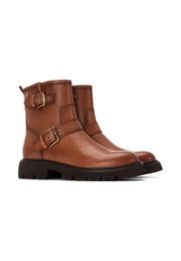 Botas de cuero marrón a la altura del tobillo con un acabado suave, que cuentan con dos hebillas metálicas decorativas y una suela de goma negra resistente.