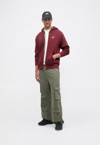 Casaco com fecho zip burgundy com bolsos frontais, calças cargo verdes-oliva e sapatilhas pretas. O modelo usa uma boné preto com um logotipo.