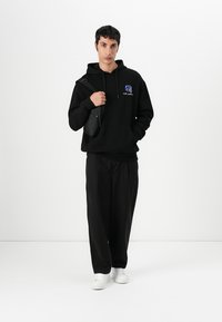 Felpa nera con cappuccio, dotata di tasca frontale e logo blu. Abbinata a pantaloni neri larghi e sneakers bianche, indossata con una piccola borsa nera.