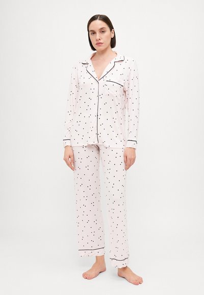 Ανοιχτό ροζ σετ πιτζάμας με εκτύπωση μαύρης καρδιάς, μαύρη επένδυση στον γιακά και την τσέπη, μπλουζάκι με μακριά μανίκια και φαρδιά παντελόνια.