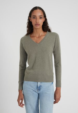 Jonge vrouw met krullend haar in een olijfgroene V-hals trui en lichtblauwe jeans, staand tegen een gewone achtergrond.