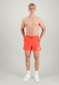 Nike úszószort fekete korall színben, rugalmas derékkal és oldalsó zsebekkel, fehér logóval. Fehér sportcipővel és zoknival viselve.