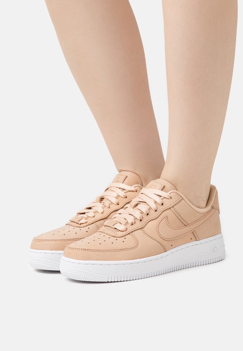 Pieds portant des baskets Nike Air Force 1 beige avec semelles blanches, lacées, sur un fond clair uni.