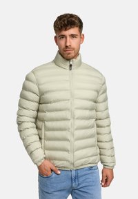 Lys grå pufferjakke med vandret syninger, høj krave og frontlynlås. Har elastiske manchetter og lommer. Båret over blå jeans.