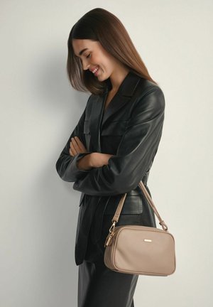 Młoda kobieta w czarnej skórzanej kurtce niesie beżową torbę na ramię, uśmiecha się i patrzy w dół, skrzyżowane ramiona na jednolitym tle.