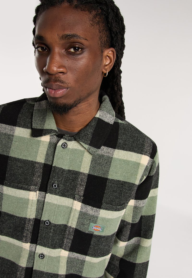 Junger Mann mit Dreadlocks, der einen goldenen Creolenohrring und ein kariertes Kurzarmhemd in grün, schwarz und creme mit Dickies-Logo-Patch trägt.