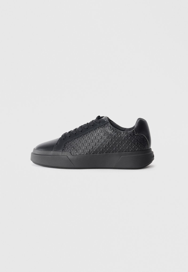 Black sneaker made of textured synthetic material, featuring a diamond pattern, round toe, flat rubber sole, and classic lace-up design.