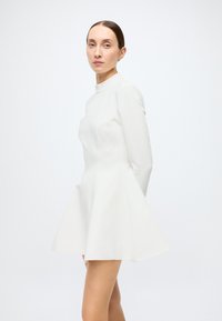 White mini dress with a high neckline, flared skirt, and long sleeves; smooth texture; minimalist design with no visible patterns or embellishments.