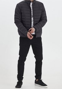 Giacca imbottita nera con cuciture verticali, colletto alto e chiusura con cerniera. Abbinata a pantaloni neri e scarpe nere; camicia bianca sotto.