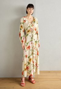 Abito maxi a stampa floreale in crema con maniche lunghe, scollatura volant e rose e fiori vibranti, abbinato a tacchi rossi con cinturini.