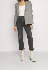 Mujer vestida con una chaqueta gris a rayas sobre una camiseta blanca sin mangas, jeans negros recortados con dobladillos desgastados y botas blancas hasta el tobillo con tacones.