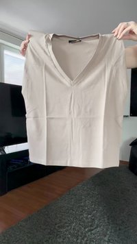 Mâini care țin un bluză scurtă cu mânecă scurtă și decolteu în V, de un bej deschis, cu un material neted și ușoare cute, în interior, lângă o fereastră și un televizor.