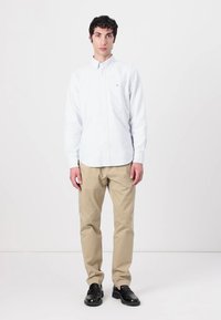 Hvid skjorte med knapper, beige bukser, sorte sko. Glat stof, tætsiddende snit og minimal design detalje. Model poserer foran en ensfarvet baggrund.