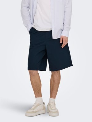 Bărbat purtând pantaloni scurți largi bleumarin, pantofi sport albi, șosete albe, un tricou alb și o cămașă deschisă la culoare cu dungi, stând cu o mână în buzunar.