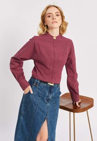 Donna che indossa un maglione bordeaux a maniche lunghe e una gonna di jeans blu alta in vita con uno spacco laterale, appoggiata a uno sgabello di legno.