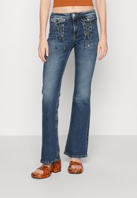 Rozšířené modré džíny z denimu s detailním šněrováním vpředu, stříbrným kováním a prošívanými akcenty. Nosit s oranžovými sandály na platformě s hadím potiskem.