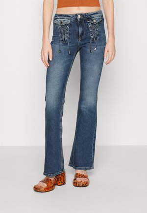 Vaqueros de mezclilla azules acampanados con detalle de cordones en la parte frontal, herrajes plateados y acentos cosidos. Llevados con sandalias gruesas de estampado de serpiente naranja.
