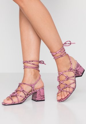 Pink højhælede sandaler med et tekstureret slangemønster, der har et stropdesign og bundet ankelaccent. Klodshæl og åben tå.