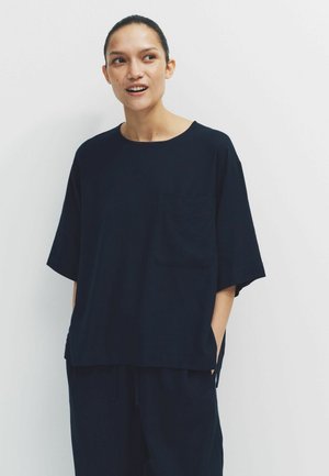 Mladić s kosom vezanom na leđima, odjeven u labavu tamno plavu kratku majicu s rukavima i hlače koje se slažu, s rukama u džepovima, gleda bočno.