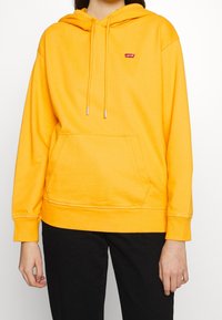 Sudadera amarilla con capucha, bolsillo frontal, cordones ajustables y un pequeño logo rojo de Levi's en el lado izquierdo del pecho. Tejido suave, corte relajado.