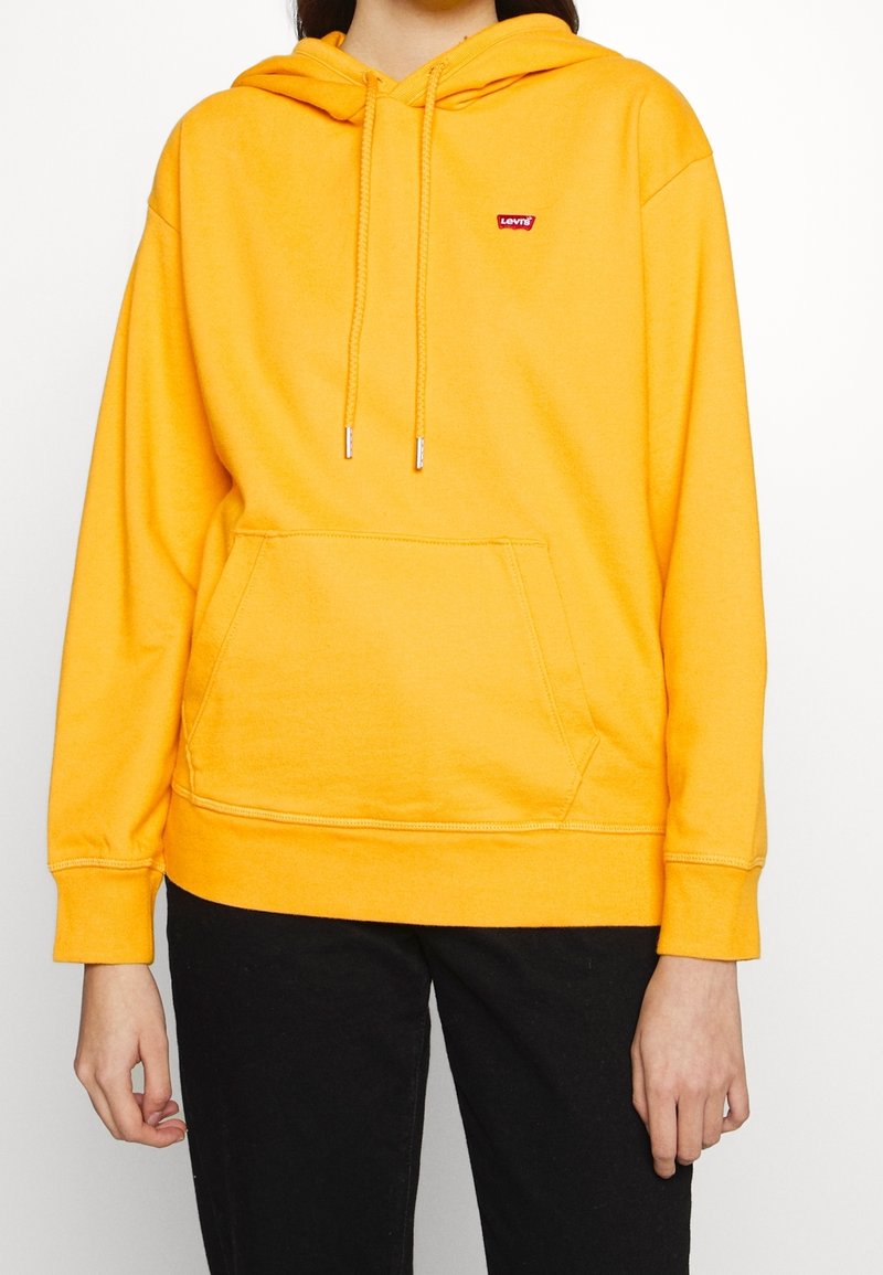 Sudadera amarilla con capucha, bolsillo frontal, cordones ajustables y un pequeño logo rojo de Levi's en el lado izquierdo del pecho. Tejido suave, corte relajado.