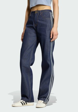 Person iført mørkeblå jeans med tre lysegrå striber ned langs siderne og hvide sneakers med sorte striber.