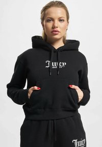 Černá mikina s kapucí, přední kapsou a bílým logem "Juicy Couture". Měkká tkanina s šňůrkami a žebrovanými manžetami. Stylová v kombinaci s odpovídajícími kalhotami.