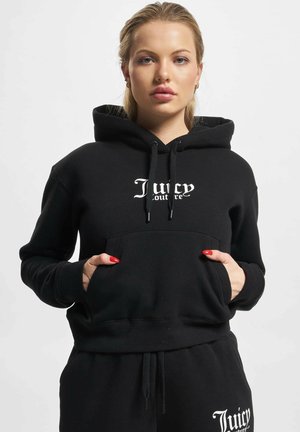 Zwarte capuchontrui met een voorzak en een wit "Juicy Couture" logo. Zachte stof met trekkoorden en geribbelde mouwen. Gestyled met bijpassende broek.