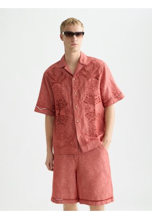 Giovane uomo che indossa una camicia rossa ricamata a maniche corte e pantaloncini coordinati, con occhiali da sole scuri, in piedi davanti a uno sfondo bianco uniforme.