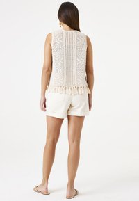 Top de punto sin mangas en color crema con un patrón y dobladillo con flecos, combinado con pantalones cortos blancos remangados. Textura suave, diseño informal.