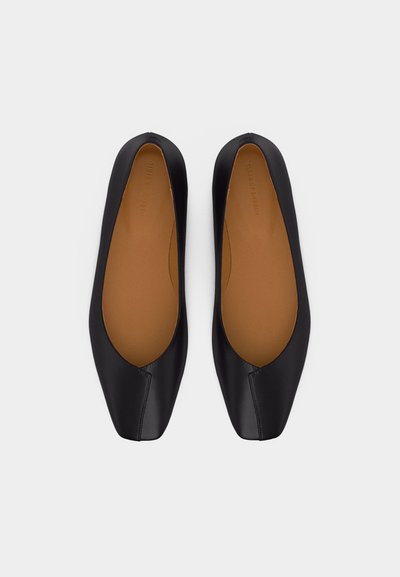 Ζευγάρι μαύρα μπαλαρίνια από δέρμα με τετράγωνα δάχτυλα και ταφ χρωματιστά πέλματα, εμφανιζόμενα από πάνω σε ανοικτό φόντο.