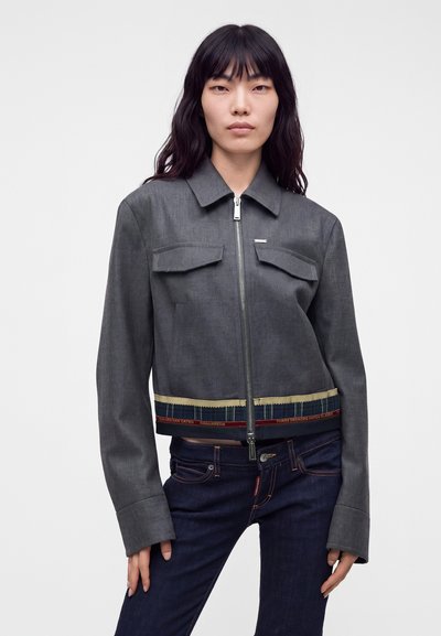 Femme portant une veste grise zippée avec des poches à rabat et un jean bleu foncé, debout devant un fond clair uni.