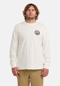 Mann mit schulterlangen Haaren, der ein weißes Langarmhemd mit einem kleinen bunten Logo auf der linken Brust und beigen Hosen trägt.