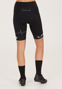Zwarte fietsshorts met een hoge tailleband, voorzien van een hartslaglijn graphic en 'ELITE LAB' branding. Gecombineerd met zwarte sokken en schoenen.