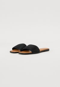 Sandales glissantes tressées noires avec une semelle plate, dotées d'un dessus texturé et d'une semelle intérieure lisse couleur tan. Conçues pour un usage décontracté.