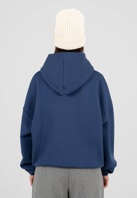 Sudadera con capucha de gran tamaño en azul marino, con cordón en la capucha, hecha de material suave. Combinada con un gorro de punto en color crema, con textura acanalada.