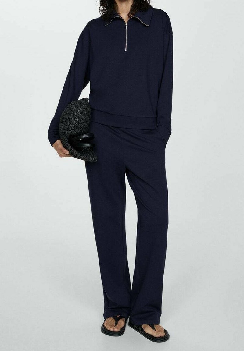 Personne portant un haut zippé bleu marine à manches longues et un pantalon assorti, tenant un chapeau noir texturé et portant des tongs noires.