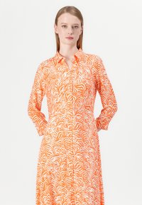 Oranssi mekko, jossa on valkoinen aaltoileva tiikeriraidallinen kuvio, pitkät hihat, kauluskainalot ja nappikiinnitys edessä.