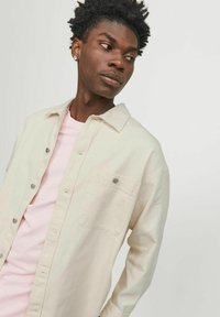 Light beige button-up shirt with a chest pocket and silver hardware paired with a soft pink crew-neck t-shirt underneath.